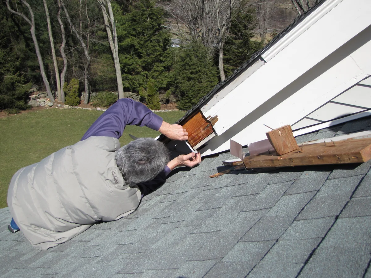 Carpenter repairing damaged soffit wood leaning onto roof shingles from a ladder